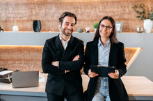 Portrait Of Two Caucasian Confident Successful Company Employees, Business Colleagues, A Man And A Woman, Stand Near The Desktop In A Modern Office, Look At The Camera, Smile Friendly