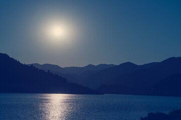 Wall Mural - Mountain lake, the silhouette of the mountains and the surface of the water, the moon, the sun reflected in the water. Beautiful blue landscape