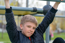 Portrait Of Confident Teen Boy In Warm Jacket Looks Directly Into Camera On Climb Equipment At Town Park.