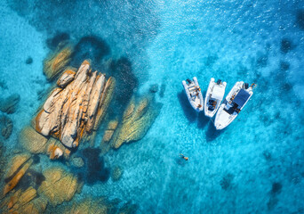 Aerial view of beautiful boats and stones in blue sea at sunset in summer. Sardinia island, Italy. Top view of yachts and rocks in transparent water. Motorboats on sea coast Travel. Tropical landscape