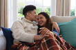 Happy biracial couple sitting on couch under blanket, laughing and embracing in living room