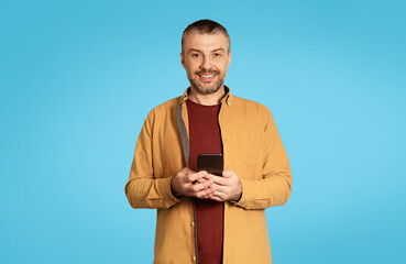Poster - Cheerful Middle Aged Male Holding Smartphone Over Blue Background