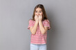 Portrait of dark haired nervous adorable little girl wearing striped T-shirt biting his fingers with shocked look, fears and phobias. Indoor studio shot isolated on gray background.