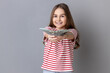Portrait of satisfied little dark haired girl wearing striped T-shirt holding out dollar bills at camera, boasting money won in lottery. Indoor studio shot isolated on gray background.
