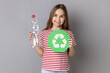Portrait of satisfied smiling little girl wearing striped T-shirt holding empty plastic bottle and recycling sign, thinking green. Indoor studio shot isolated on gray background.
