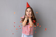Portrait of happy little girl wearing striped T-shirt and party cone blowing heart shaped confetti, enjoying birthday, festive mood. Indoor studio shot isolated on gray background.