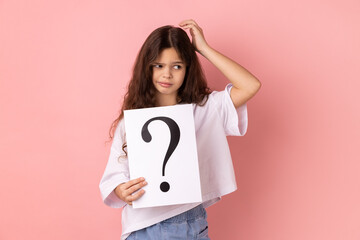 Portrait of little girl wearing white T-shirt holding paper with question mark over, thoughtful, face thinking about question, very confused idea. Indoor studio shot isolated on pink background.