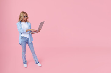 Portrait of a young girl in casual denim blue clothes with a laptop in her hands on a pink background. The concept of learning and job search online