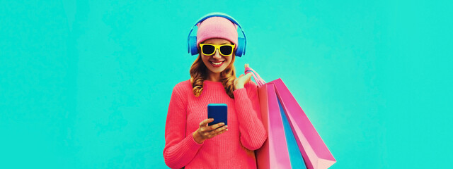 Colorful portrait of stylish smiling young woman listening to music in headphones with shopping bags posing wearing knitted sweater, pink hat on blue background