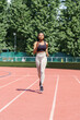 © DimaBerlin - Focused sportive black woman enjoys training on sunny summer day preparing for upcoming marathon. Young African American athlete runs on red track of stadium against city park with lush green trees