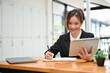 © bongkarn - Asian businesswoman at her desk, using tablet and working on report.