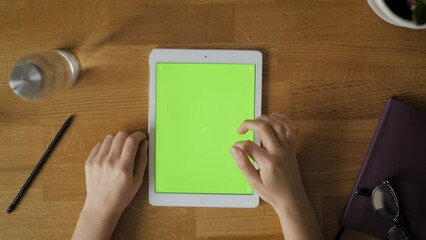 Overhead shot of tablet with green screen lying on the wooden table and person using it. Touching, swiping, browsing.