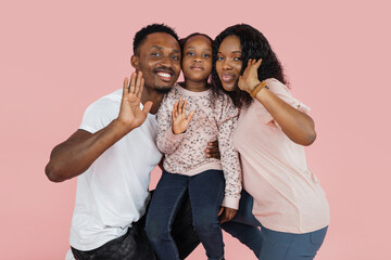 Wall Mural - Close up portrait of excited african american man, woman and girl laughing and posing isolated on pink studio wall. Cheerful father hugging daughter and wife waving their hands .