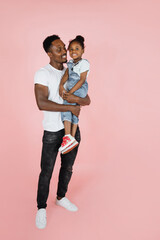 Wall Mural - Family love. Vertical portrait of happy afro daddy embracing with his cute baby daughter, pink studio background.