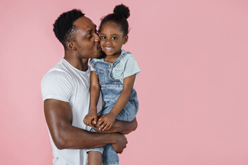 Wall Mural - Family love. Horizontal portrait of happy afro daddy embracing with his cute baby daughter and kissing her in cheek, pink studio background.