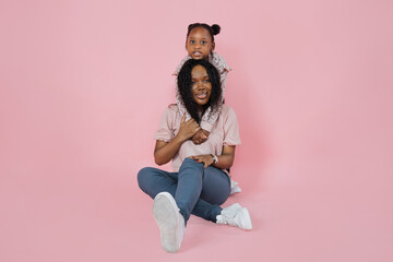 Wall Mural - Fun with mom, happy childhood. Pretty African American mother sitting on floor and her daughter hugging her from back, smiling at camera. Isolated pink background with copy space.