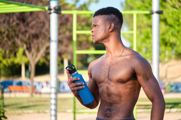 Wall Mural - Portrait of a young fit shirtless black man holding water bottle looking at a side in a park outdoors. Fitness and sport lifestyle.