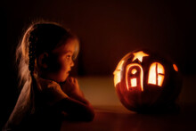 Happy Excited Little Girl Looking Inside Of Glowing Jack O Lantern, Carved Pumpkin, While Standing In Dark House Room Decorated For All Hallows Eve. Halloween Day, Selective Focus
