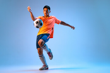 Portrait of young man, football player in motion, action, kicking ball with knee isolated over blue studio background in neon light