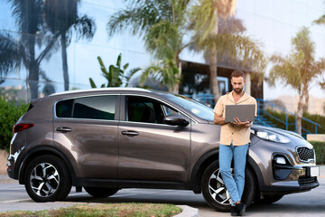 Pensive middle aged latin man using laptop computer, working online standing near new car. Handsome driver planning road trip  