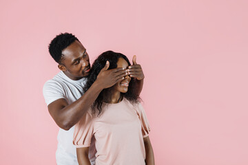 Wall Mural - Cheerful funny young african couple friends man woman in casual clothes, male close eyes of his girlfriend with hands, playing guess who, or hide and seek isolated on pink background studio portrait.