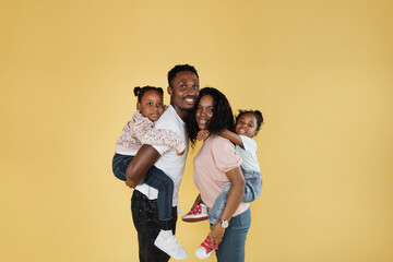 Wall Mural - Time with family. Overjoyed african american family laughing and posing isolated over yellow studio wall. Cheerful father and mom carrying their daughters on their back.