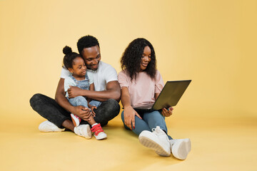 Wall Mural - Happy family, technology, communication concept. Young african parents sitting on floor with their daughter and embracing, using laptop for video call, over yellow studio background, free space.