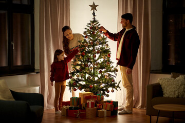 Poster - family, winter holidays and people concept - happy mother, father and little daughter decorating christmas tree at home