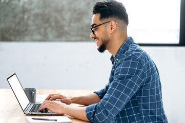 White laptop screen, mockup concept. Arabian or Indian young man, stylishly modernly dressed, programmer, freelancer, sitting at the table with a laptop, typing on the keyboard