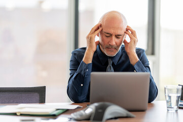 Wall Mural - Stressed middle aged businessman having a migraine while sitting at office desk and working on laptop