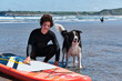 Young surfer with long curly hair posing with his border collie dog and his paddle surf board
