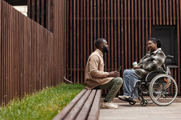 Graphic side view shot of young woman in wheelchair talking to friend or colleague outdoors in city park, copy space