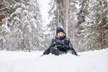 Wall Mural - Happy teenager boy sitting on snow in winter forest. Child having fun outdoors. Joyful adolescent playing in snow at snowfall. Laughing smiling kid walking in winter park in cold weather