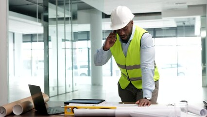 Poster - A male engineer talking on the phone, happy and smiling in an office, at a desk with blueprints and equipment. A successful African architect on a call confirming designs for a construction project.