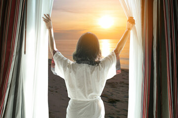 Wall Mural - The woman is on the window in the bedroom. She opened the curtain at the window. In the morning and she looked at the view of the sea beach during the sunrise time