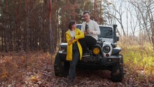 Young Lovely Couple With A White Car In Autumn Forest. They Drinks Tea From The Thermos