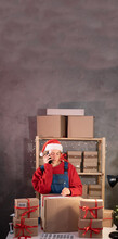 Woman In Santa Claus Hat Works In A Warehouse Of Boxes With Gifts And Orders From An Online Store For Christmas. Small Business Owner Sells Happy New Year. Delivery Holiday. Banner