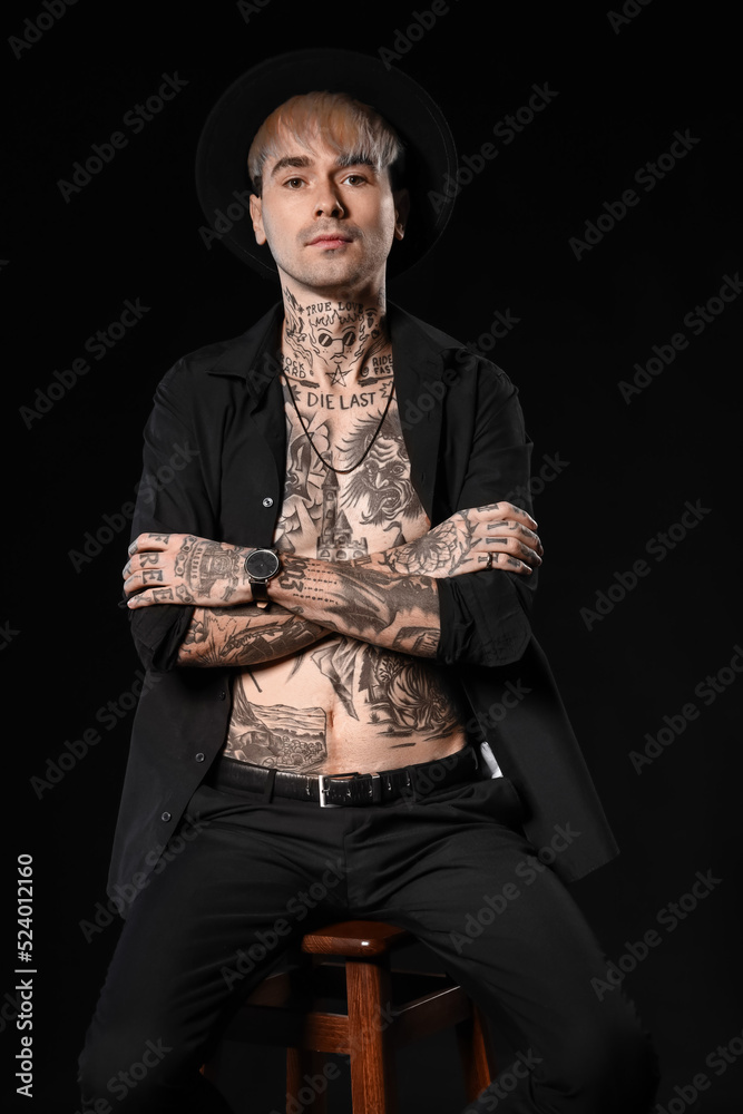 Young tattooed man sitting on stool against black background
