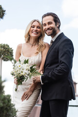 Positive newlyweds with bouquet looking at camera on terrace.