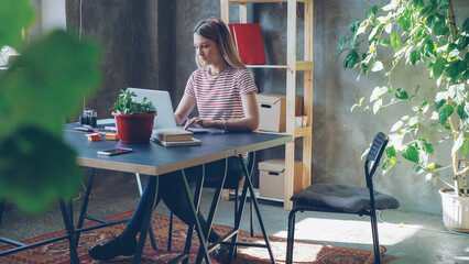 Young blond woman is working with laptop sitting at table in office. Her colleague is coming, women start watching screen together and laughing. Informal friendly atmosphere.