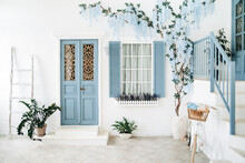 Exterior Of A White Mediterranean-style House With A Blue Door And A Window, A Flowering Tree. Traditional Patio Of Santorini