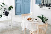 Romantic Seating Area With Beige Chairs And A Vintage Table With Two Cups Of Coffee And A Croissant On A Blurred Background Of Blue Door Of A Mediterranean White House