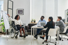 African Businesswoman In Wheelchair Pointing At Flipchart And Presenting Her Report To Colleagues During Teamwork At Office