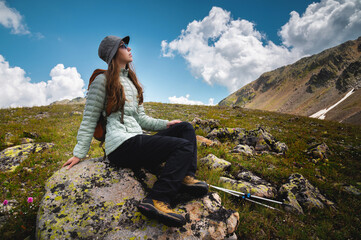 a young woman with a backpack sits on a stone and looks at the sky with clouds and mountains, hiking