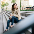 © contrastwerkstatt - Young woman with cell phone sits on stairs and looks sideways smiling