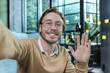 © Liubomir - Man working in modern office, happy blond businessman smiling and looking at smartphone camera, holding hand up greeting gesture, worker in glasses taking selfie, video call with friends