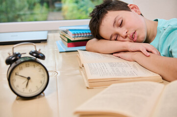 Blurred black alarm clock on the background of a tired sleepy schoolboy falling asleep on a textbook while doing homework. Teenage boy having learning disability. Back to school