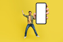 Full Body Length Portrait Of Excited Man Jumping, Making Winner Gesture And Showing Cellphone With Empty Space, Mockup