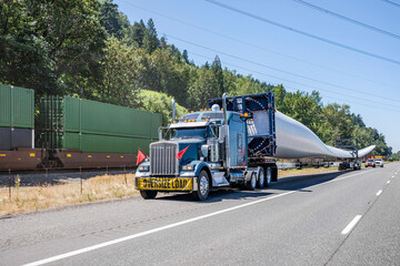 Wall Mural - Big rig semi truck tractor with oversize load sign on the front transporting windmill electric generator blade with special additional trolley standing on the highway shoulder with escort vehicles