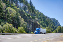 Industrial Blue Bonnet Big Rig Semi Truck With Dry Van Semi Trailer Running On The Beautiful Highway Road With Forest On The Rocky Mountain In Columbia River Gorge